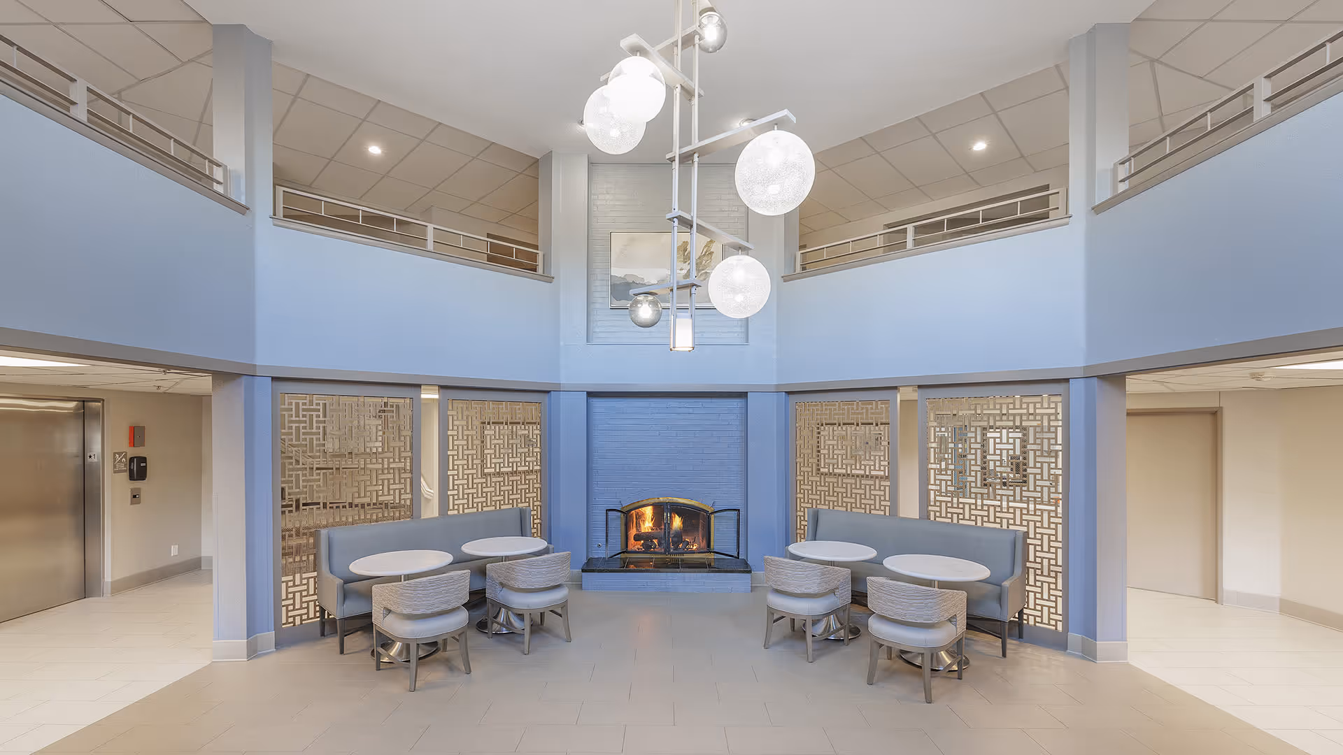 A spacious common area with a lit fireplace in the center, flanked by two seating areas each with a curved bench and round tables with chairs. The walls are painted light blue, and there is a modern chandelier with spherical lights hanging from the ceiling. The area has an open second-floor balcony overlooking the space.