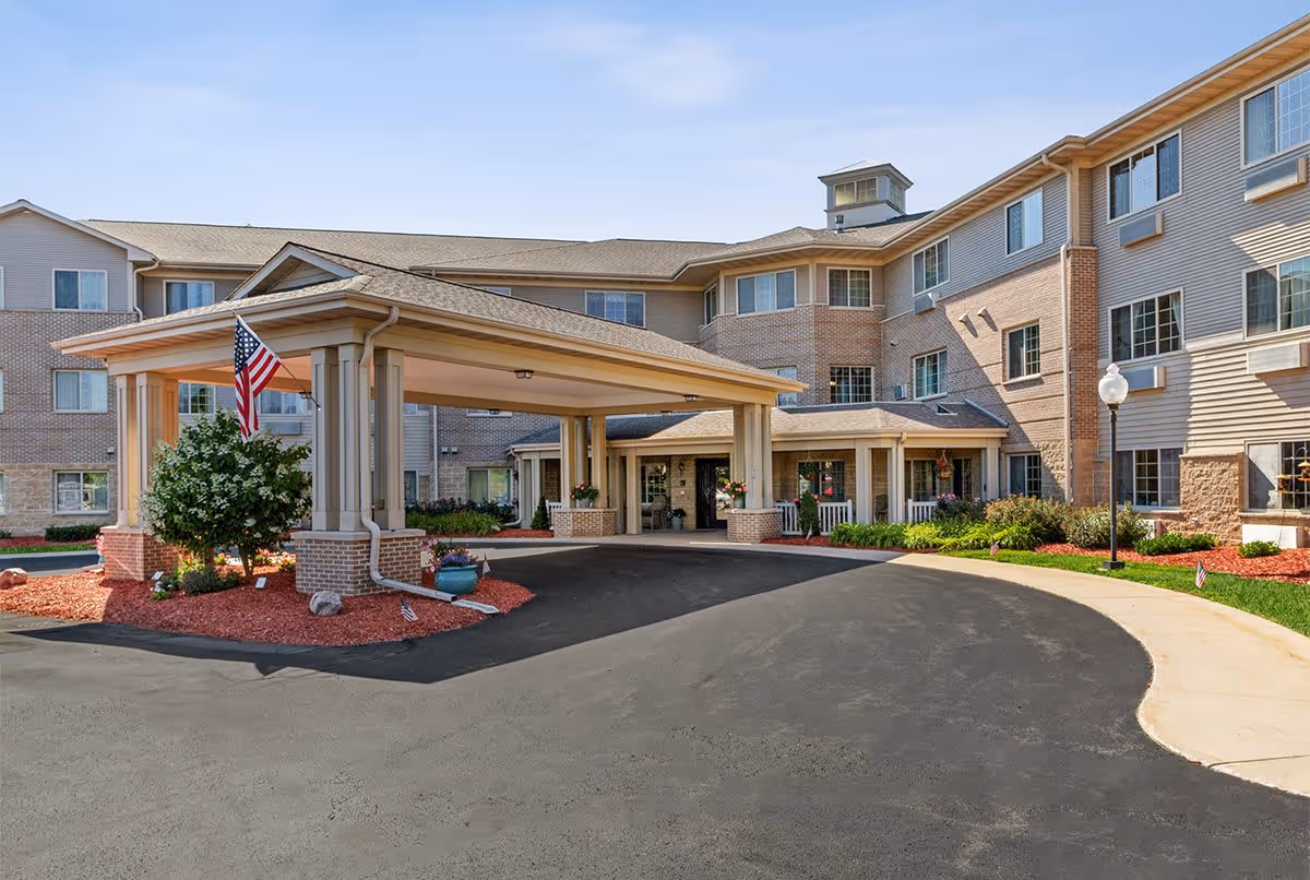 Exterior view of a multi-story senior living facility with a covered entrance driveway. The building features brick and siding exterior walls, multiple windows, and landscaped areas with bushes and mulch. An American flag is displayed near the entrance.