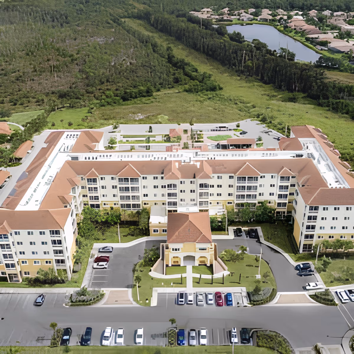 Aerial view of a large senior living facility named Discovery Village At The Forum, showing a multi-story building with a U-shaped layout, surrounded by parking lots, greenery, and a natural landscape with trees and a pond in the background.