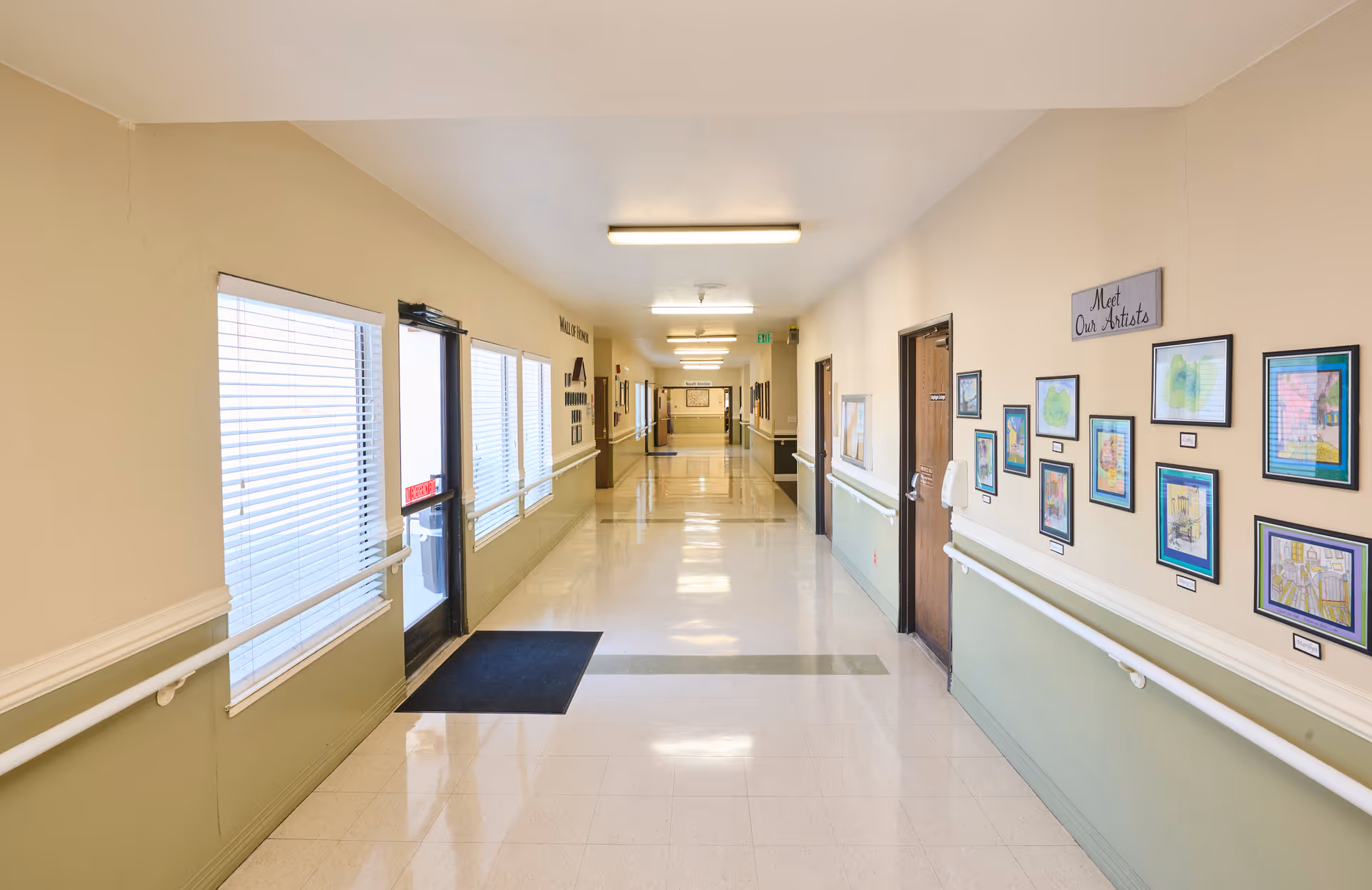 Bright, clean nursing home hallway with handrails, framed artwork along the walls, windows and an exit door at the left.