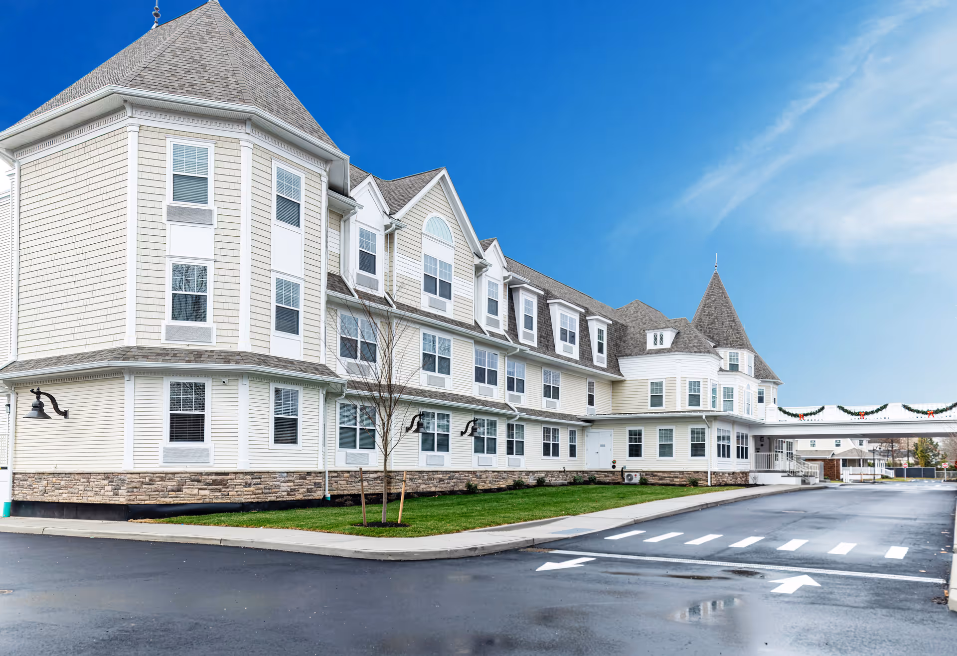 Exterior view of a large, multi-story senior living facility building with beige siding, multiple windows, and a steeply pitched roof under a clear blue sky. The building has a stone base and a driveway with directional arrows and a crosswalk in front. There are small trees planted near the building and holiday garlands hanging on a connecting walkway.