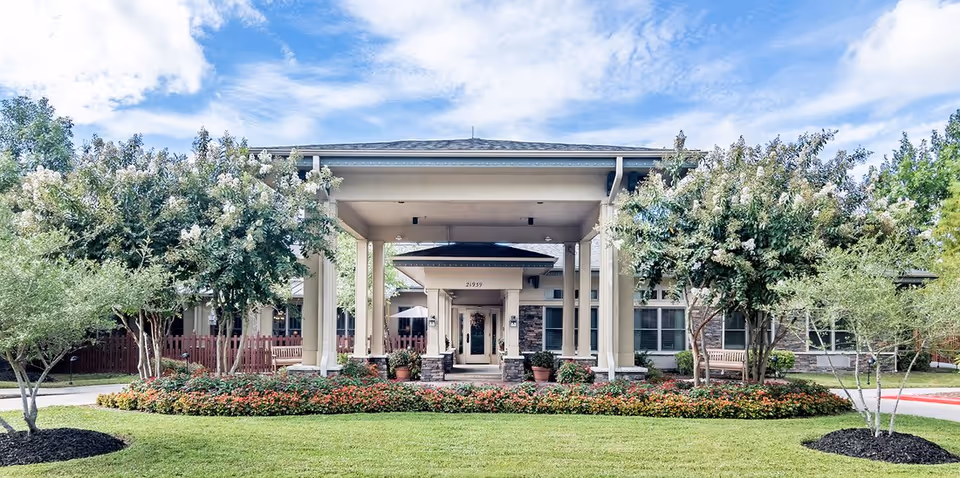 Front entrance of a single-story senior living building with a covered porte-cochere, columns, landscaped flowerbeds and trees.