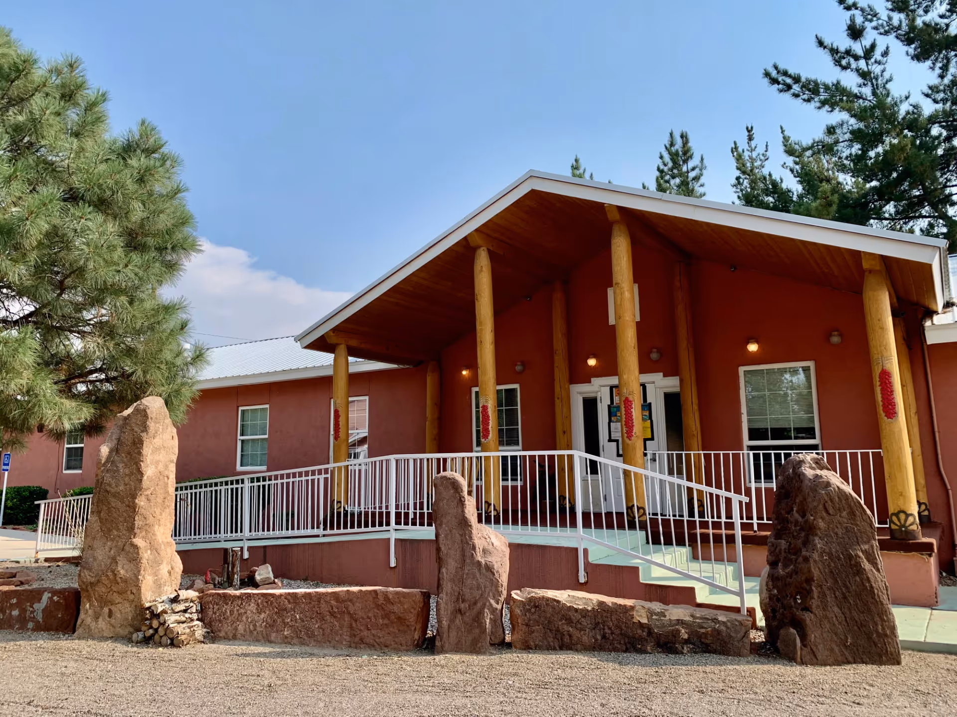 Front entrance of a single-story assisted living building with a covered porch supported by wooden posts, a wheelchair ramp, and large decorative rocks in front.