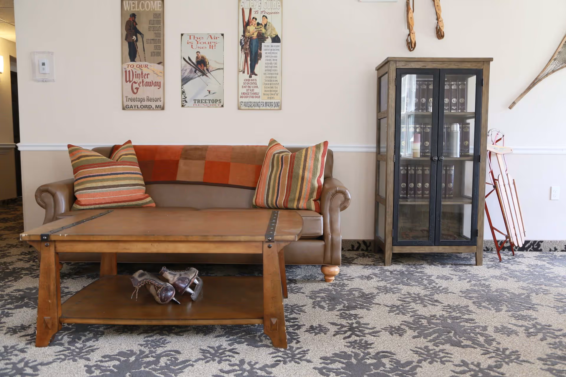 A cozy living room area with a brown leather sofa adorned with two striped pillows and a red and brown checkered throw blanket. In front of the sofa is a wooden coffee table with a pair of vintage ice skates on the lower shelf. On the wall above the sofa are three vintage-style posters. To the right of the sofa is a glass-front cabinet filled with books and other items, and a vintage sled leaning against the wall. The floor is carpeted with a patterned design.