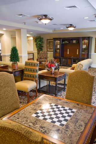 A cozy common area in an assisted living facility featuring a table with a built-in chessboard in the foreground, surrounded by upholstered chairs. In the background, there are more chairs, a wooden entertainment cabinet, decorative plants, and framed artwork on the walls. The room has warm lighting and a patterned carpet.