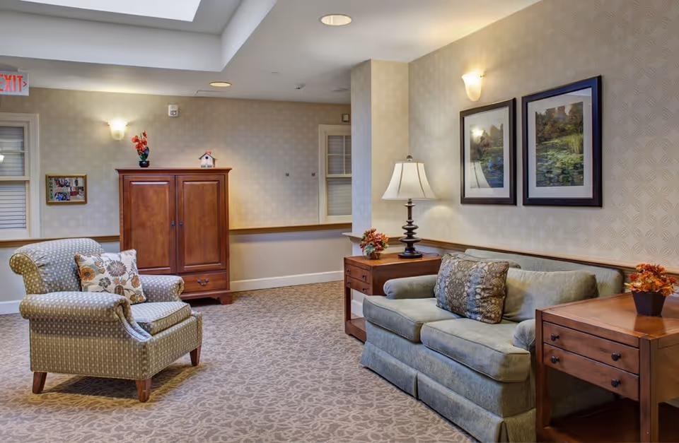A cozy living room area in a senior living facility with a patterned armchair and a floral pillow, a green sofa with two decorative pillows, two wooden side tables with lamps and small flower arrangements, a wooden cabinet against the wall, framed landscape paintings, and soft lighting from wall sconces and ceiling lights.