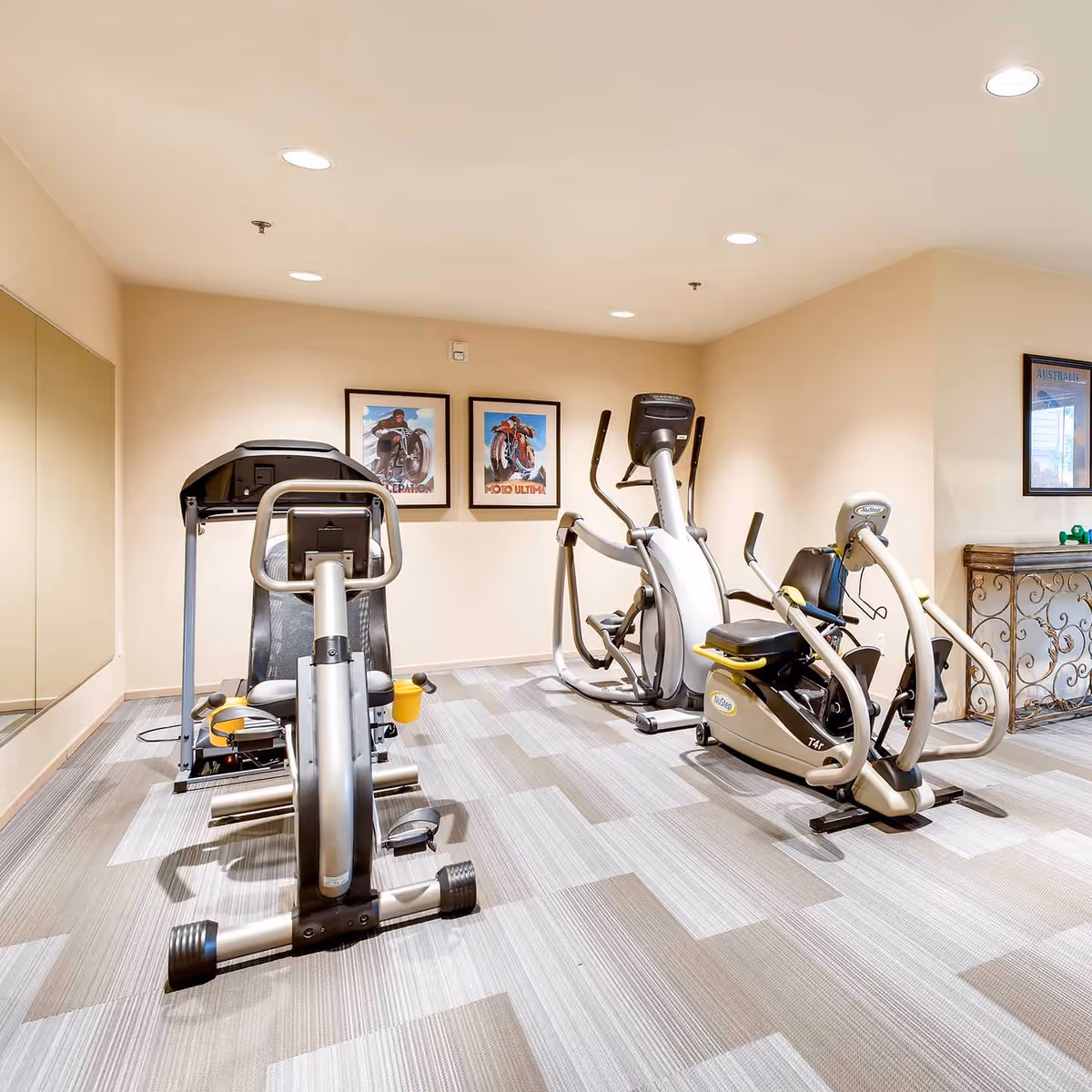 A small fitness room with three exercise machines including a recumbent bike, an elliptical trainer, and a seated upper body ergometer. The room has beige walls, a patterned carpet floor, recessed ceiling lights, a large mirror on one wall, and framed pictures of motorcycles on the wall.