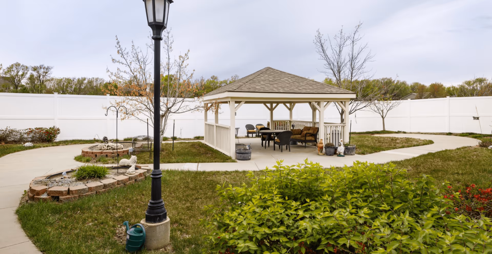 Outdoor garden area with a white gazebo featuring seating and tables, surrounded by a paved walkway, green grass, small trees, shrubs, and a white privacy fence in the background under a cloudy sky.