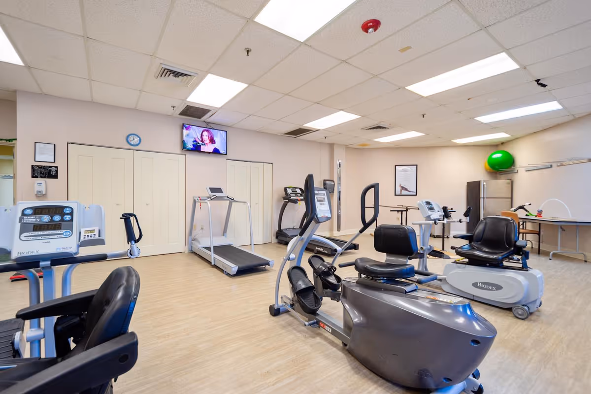 A fitness room with exercise equipment including stationary bikes, treadmills, and other cardio machines. The room has a light wood floor, beige walls, a wall-mounted TV, and a green exercise ball in the corner.