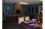 Communal dining room with several tables covered in purple tablecloths, wooden chairs, a brick fireplace, and a wall clock.