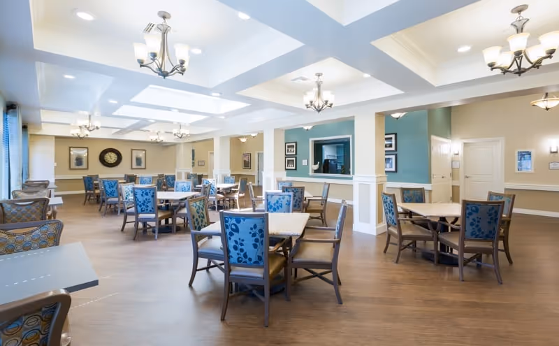 Bright, spacious dining room with multiple tables and blue-upholstered chairs under coffered ceilings and chandeliers.