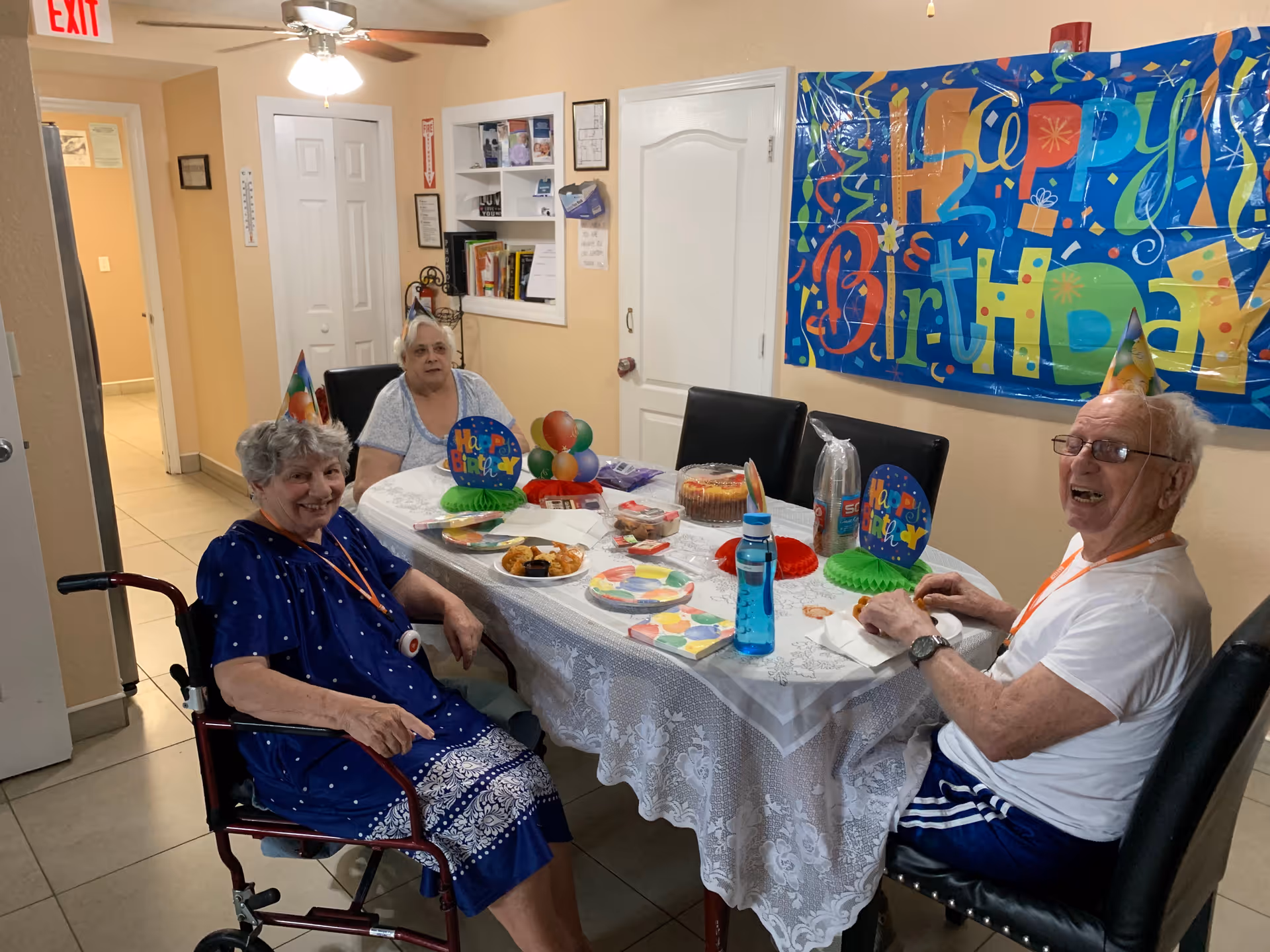 Three elderly residents sit around a decorated table celebrating a birthday in a dining area.