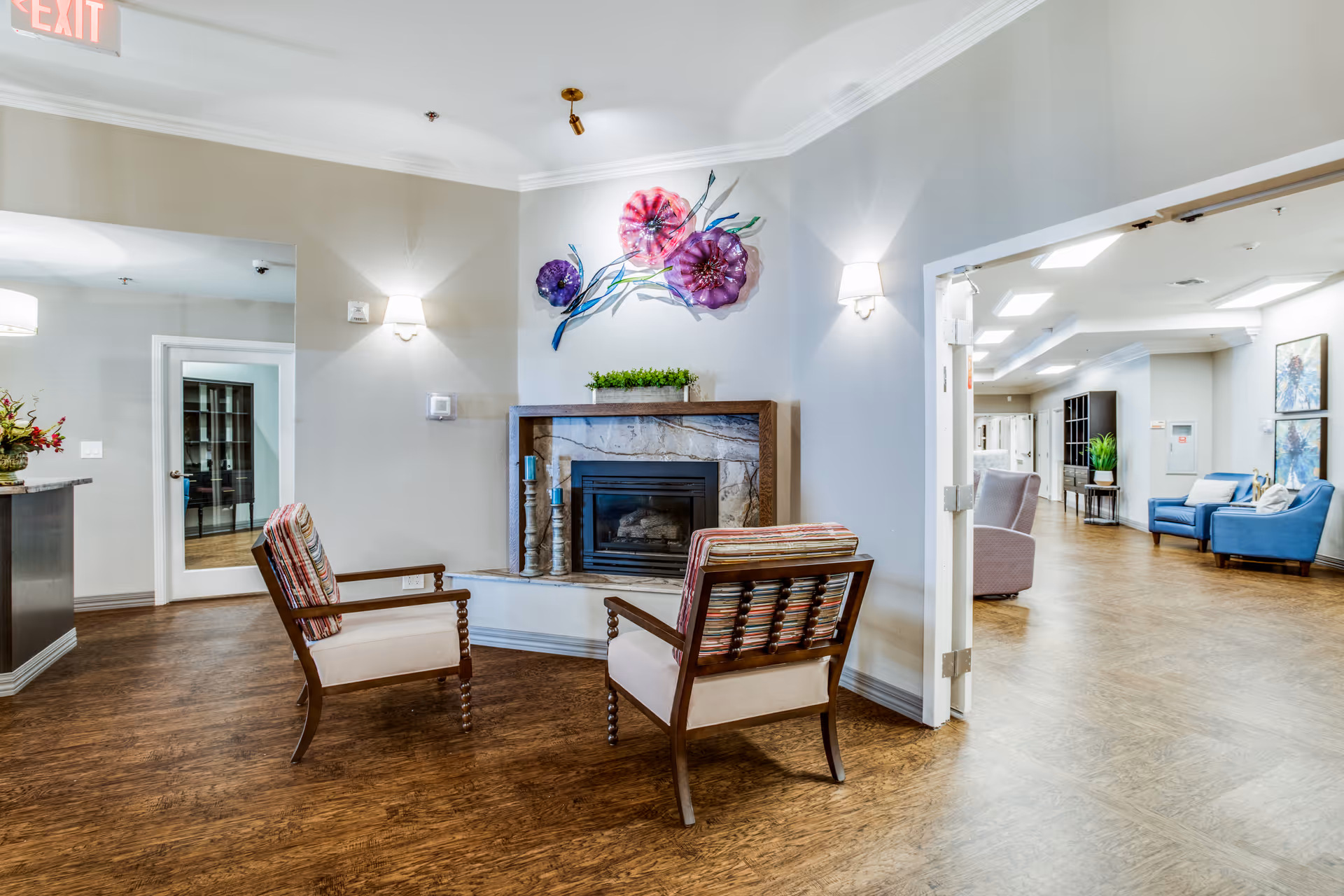 Bright communal lounge with two armchairs facing a fireplace and colorful floral wall art.
