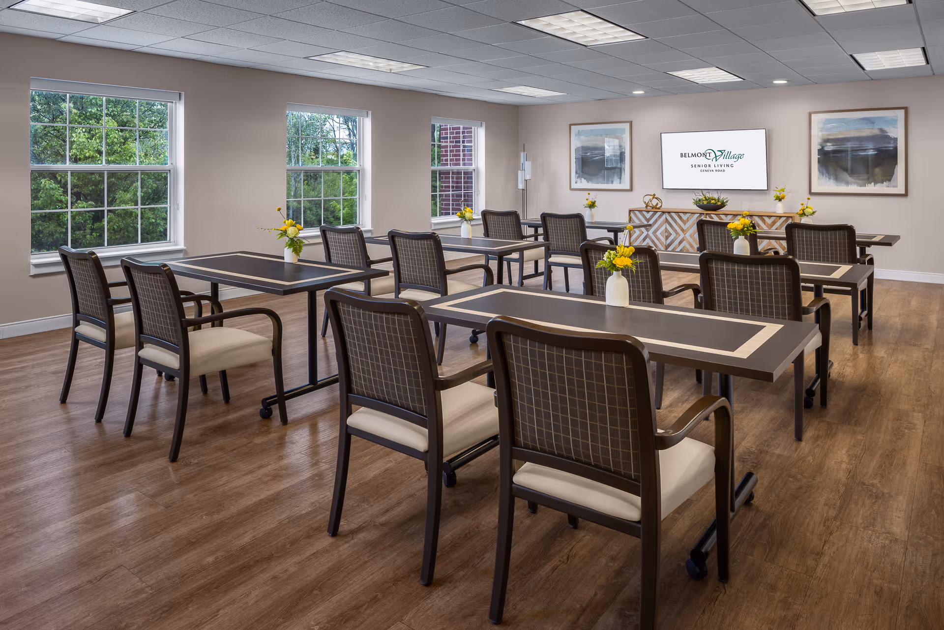 A bright and clean dining room with several rectangular tables and cushioned chairs arranged neatly. Each table has a small vase with yellow flowers. Large windows on the left wall let in natural light and show green trees outside. On the far wall, there is a flat screen displaying the Belmont Village Senior Living logo, flanked by two framed abstract paintings. The floor is wood, and the walls are painted a soft beige color.