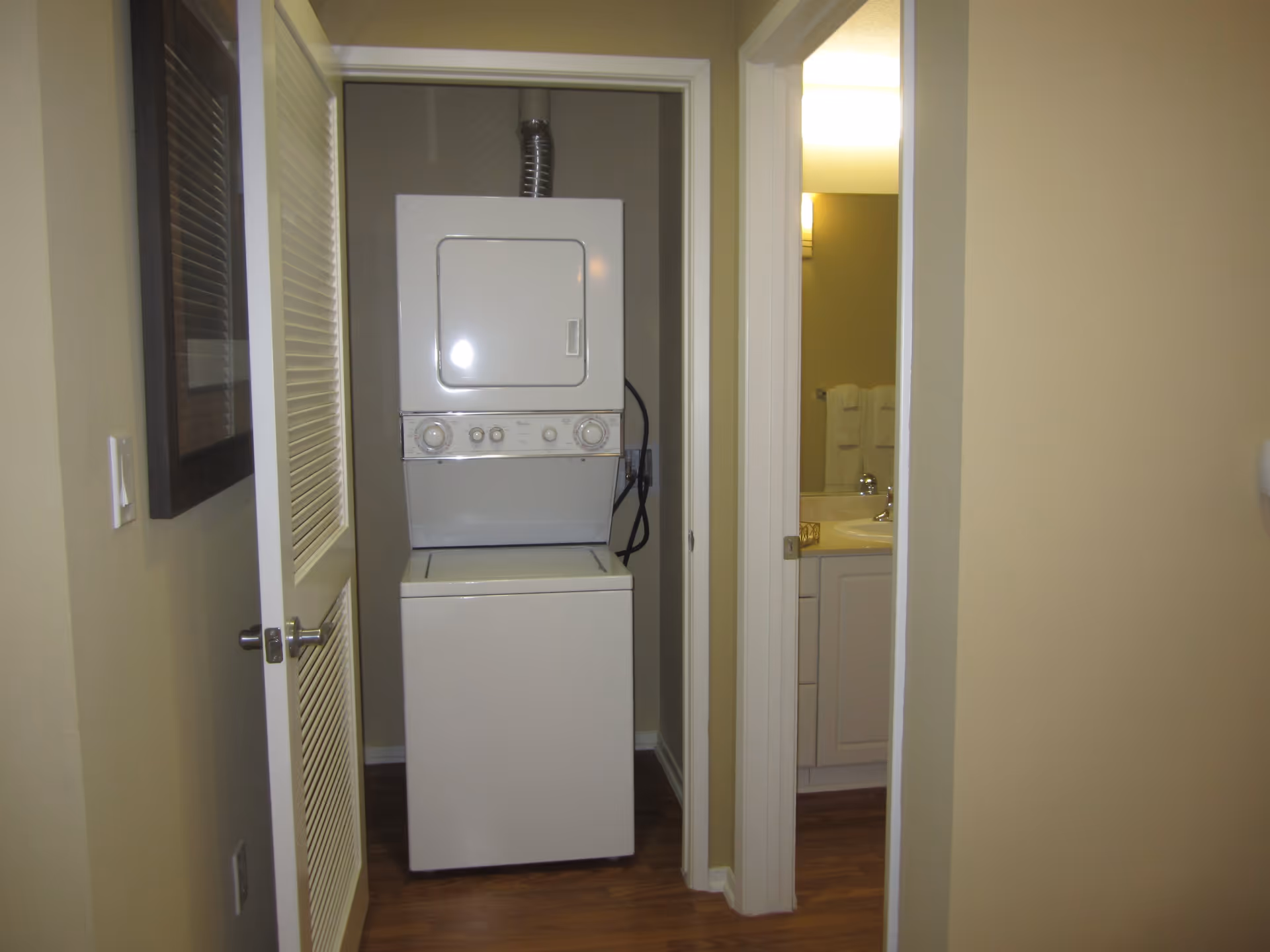 A small laundry area with a stacked washer and dryer unit inside a closet with a louvered door. To the right, there is a partially open door leading to a bathroom with a sink, faucet, and towels visible.