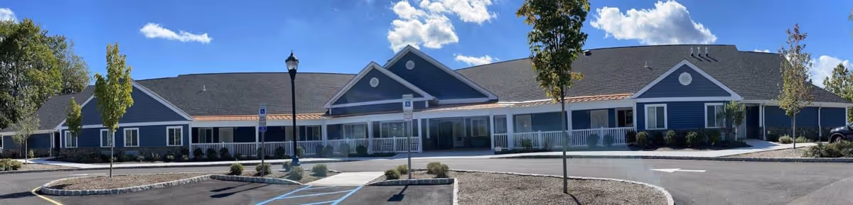 Wide panoramic view of a single-story senior living facility building with blue siding, white trim, and a gray roof under a bright blue sky with scattered clouds. The building features a covered porch with white railings, several windows, and a few small trees and shrubs in front. There are parking spaces, including handicapped spots, and a streetlamp in the paved area in front of the building.