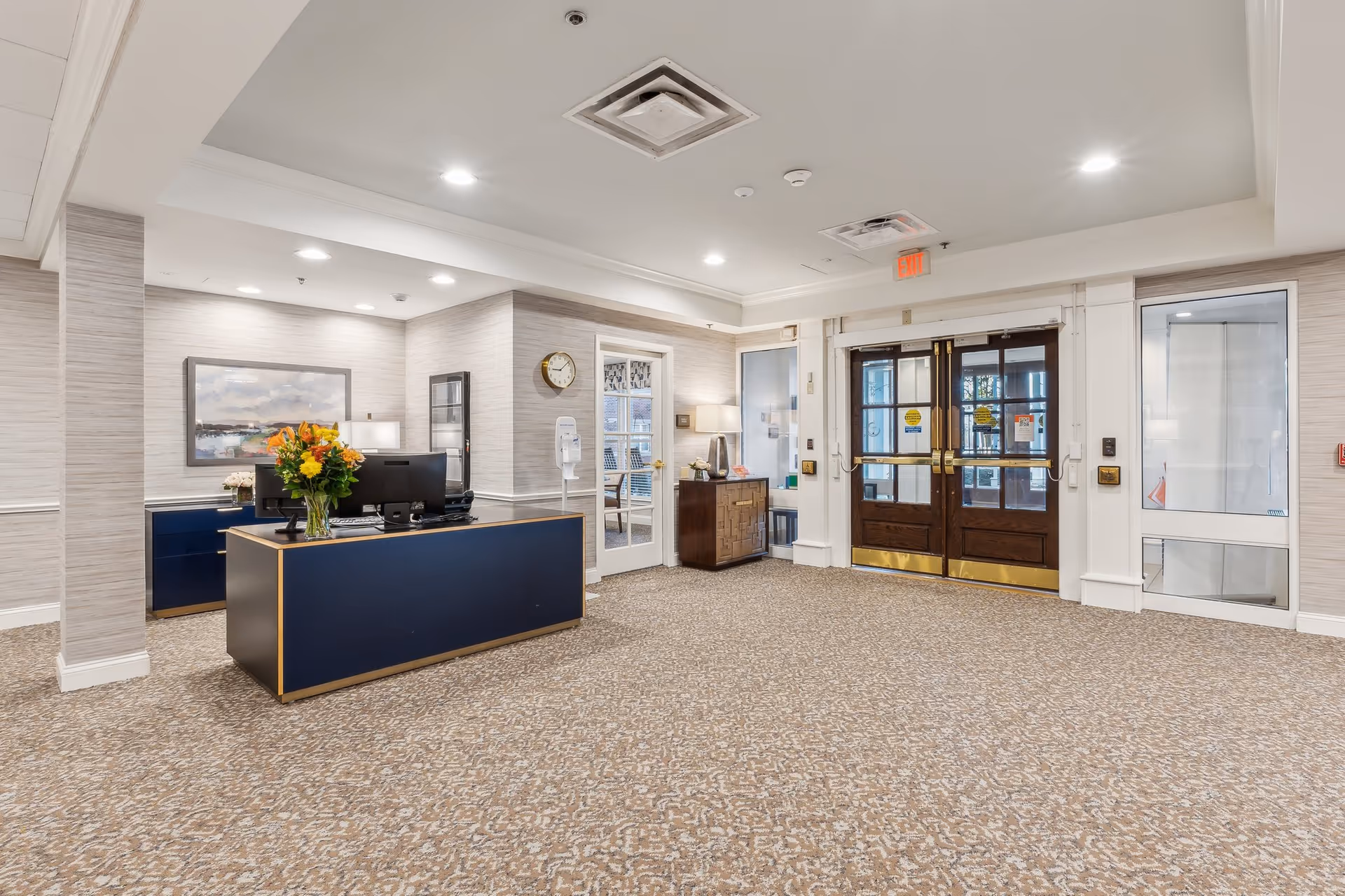 Reception area of a senior living facility with a blue desk adorned with a vase of flowers, a clock on the wall, a painting, and double wooden doors with glass panels leading outside.