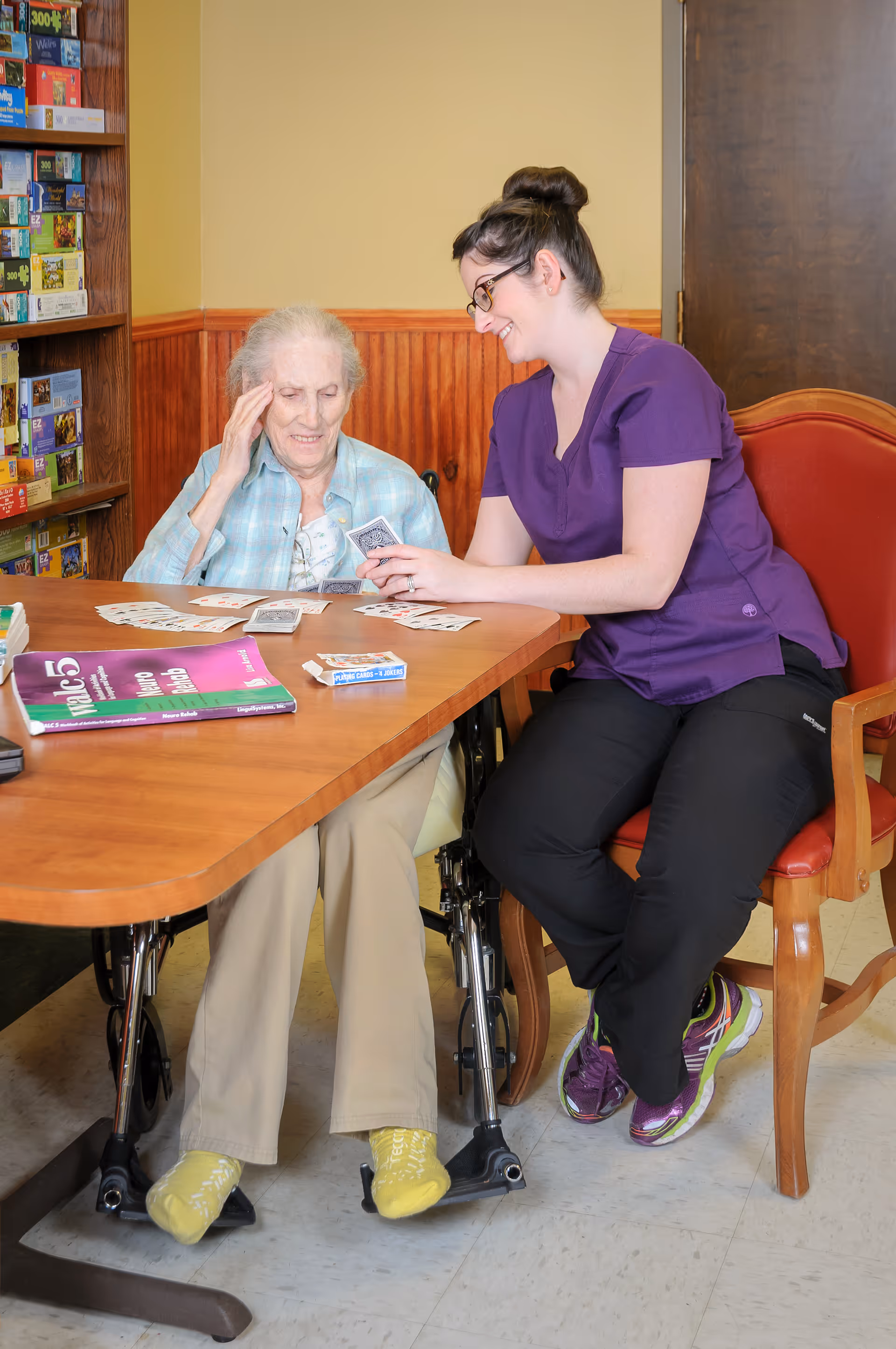 An elderly woman in a wheelchair and a caregiver in purple scrubs are sitting at a wooden table playing cards together. The elderly woman is holding her head with one hand and looking at the cards on the table, while the caregiver is smiling and holding a card. There are board games on a shelf in the background and a magazine on the table.