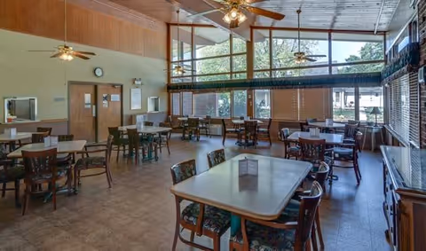 A spacious dining room with multiple tables and chairs arranged neatly. The room features large windows allowing natural light to fill the space, ceiling fans, and a high ceiling with wood paneling. There are doors and a clock on one wall, and the room has a warm, inviting atmosphere.