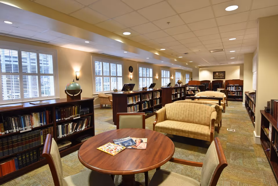 A well-lit library or reading room with multiple bookshelves filled with books, several comfortable beige upholstered sofas, and a round wooden table with chairs. The room has large windows with white shutters allowing natural light to enter, and the ceiling has recessed lighting.
