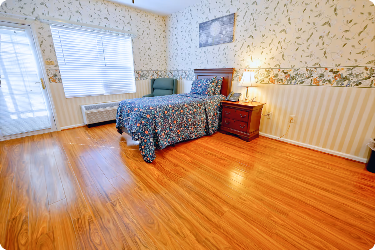 A cozy bedroom with a single bed covered in a floral patterned bedspread, a wooden nightstand with a lamp and a phone, a green armchair, floral and striped wallpaper, a window with blinds, and a door with a glass panel. The floor is wooden with a polished finish.