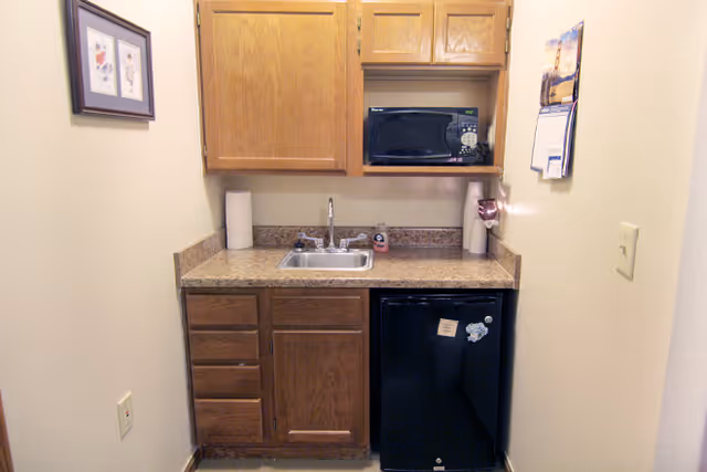 A small kitchenette area with wooden cabinets, a countertop with a sink, a microwave oven on an upper shelf, and a mini refrigerator below the counter. There are paper towel rolls on the counter, a soap dispenser near the sink, and a calendar hanging on the wall to the right. Two framed pictures are hung on the left wall.