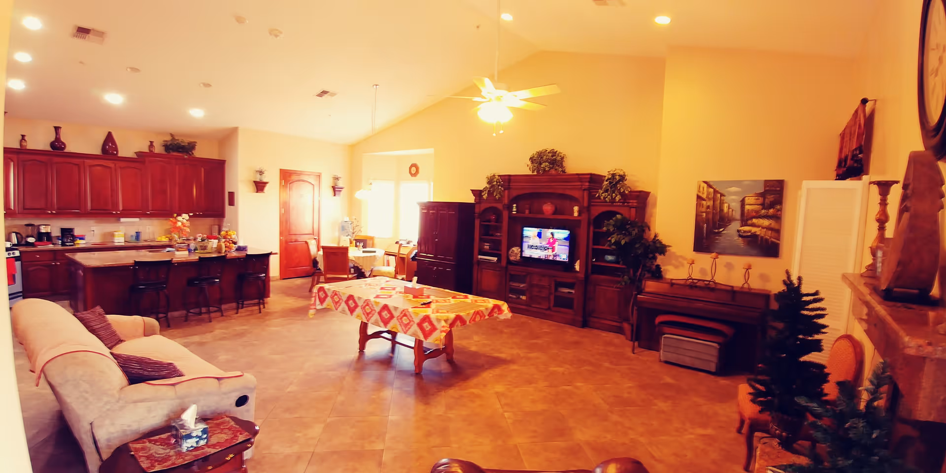 A spacious and warmly lit common area featuring a beige sofa with pillows, a wooden coffee table with a colorful tablecloth, a large wooden entertainment center with a TV, and a kitchen area with wooden cabinets and a breakfast bar with stools. The room has tiled floors, a ceiling fan with lights, and various decorative plants and wall art.