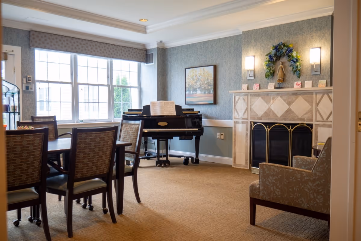 Well-lit common room with a dining table and chairs, a piano by the window, and a fireplace decorated with a wreath.