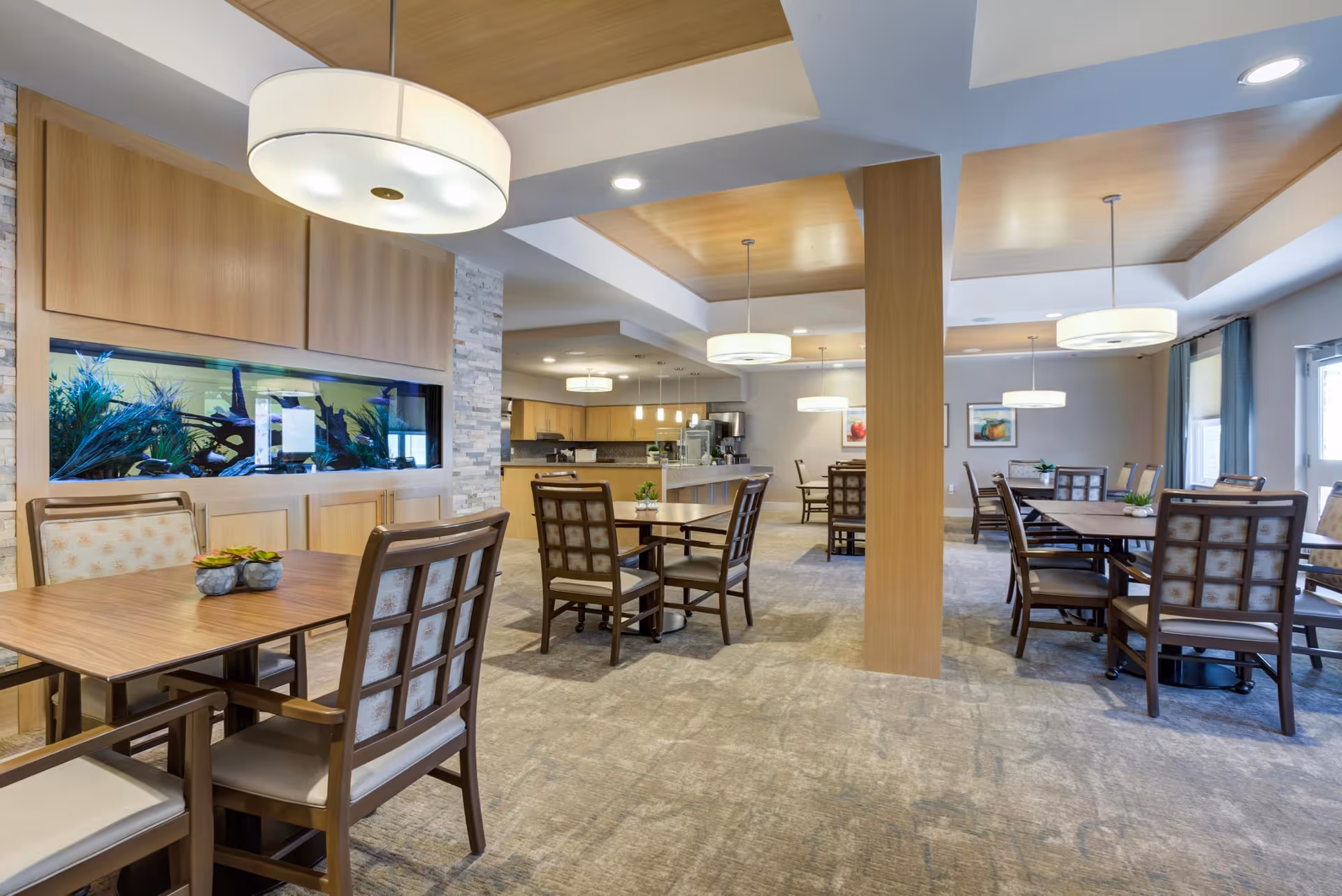 A spacious dining area with multiple wooden tables and cushioned chairs arranged neatly. The room features modern pendant lights hanging from a ceiling with wooden panel accents. There is a large built-in aquarium with plants and fish on one wall, and the flooring is carpeted in a neutral tone. The space has a warm and inviting atmosphere with natural light coming through windows on the right side.