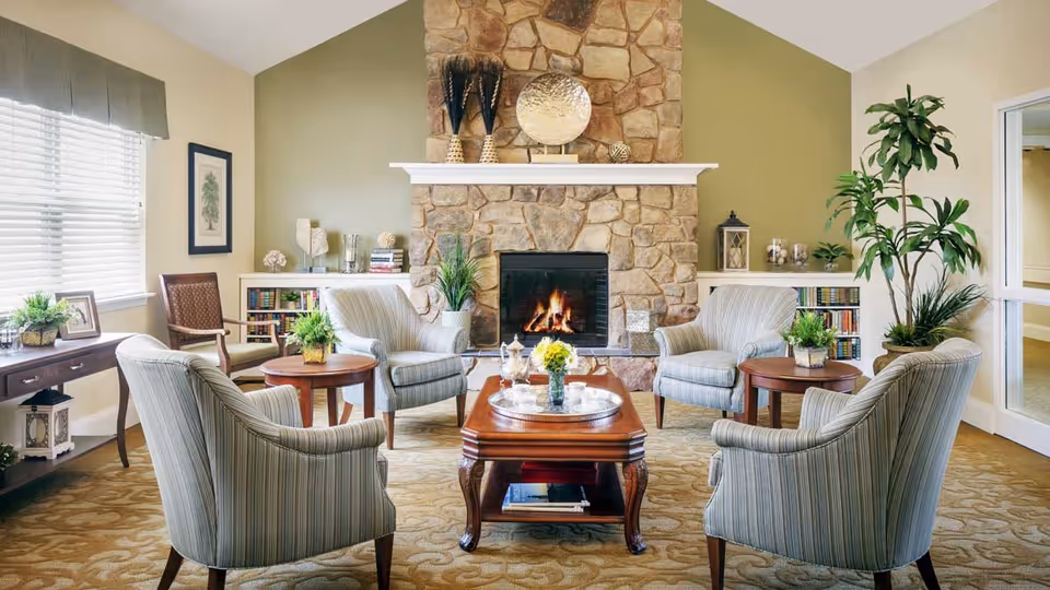 A cozy living room with four striped armchairs arranged around a wooden coffee table with a silver tray and flowers. The room features a stone fireplace with a fire burning, decorative items on the mantel, bookshelves on either side, a large window with blinds, and several green plants.