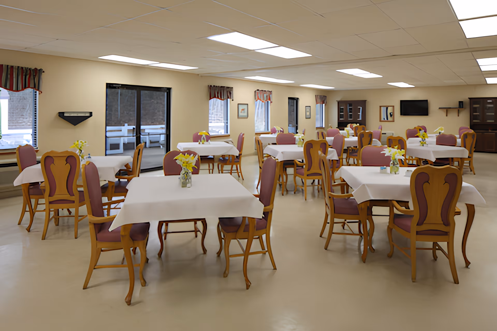 Spacious dining room with several tables covered in white tablecloths and wooden chairs, each table decorated with a small vase of flowers.