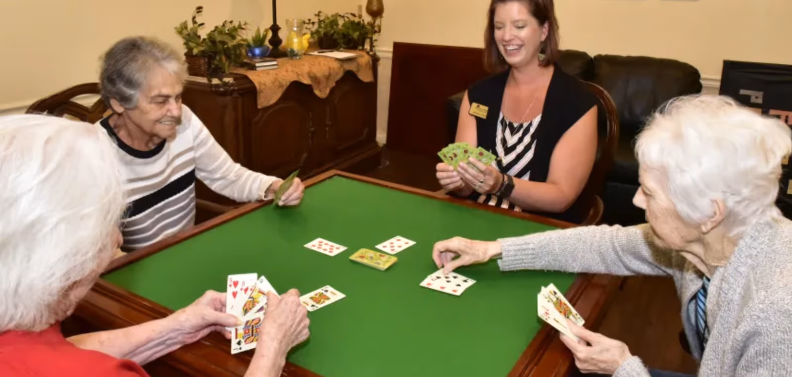 Four elderly women and a younger woman sitting around a green card table playing a card game in a cozy room with wooden furniture and plants in the background.