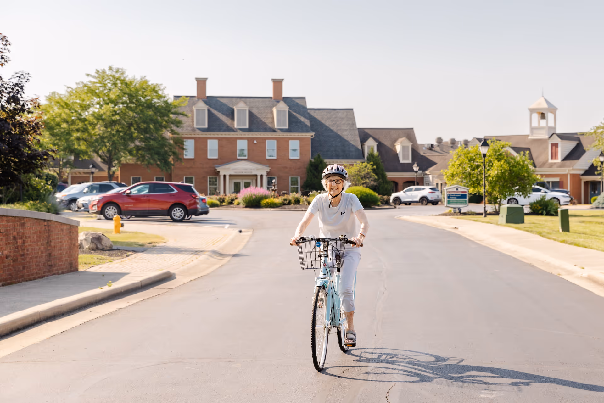 An elderly woman wearing a helmet rides a bicycle on a paved road in front of a large brick building with multiple windows and a peaked roof. Several cars are parked along the sides of the road, and there are trees and landscaped bushes around the area under a clear sky.