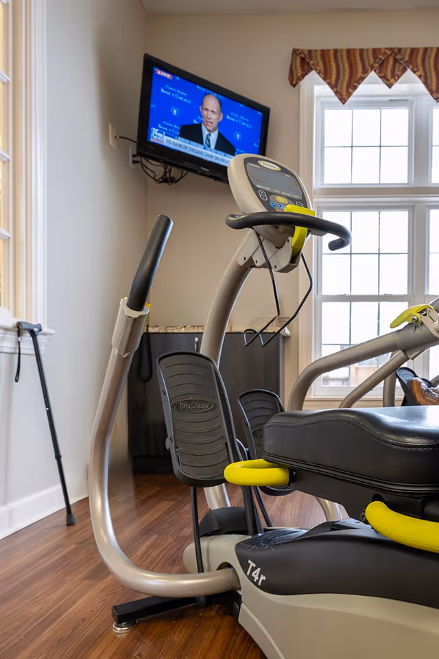 Exercise equipment including a recumbent bike and elliptical machine in a room with wood flooring, a window with a valance, a wall-mounted TV showing a news broadcast, and a walking cane leaning against the wall.