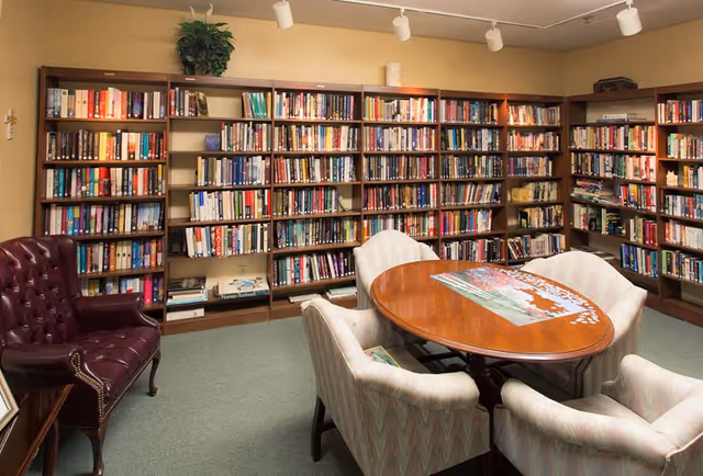 A cozy library room with multiple wooden bookshelves filled with books along the walls. In the center, there is an oval wooden table with a partially completed jigsaw puzzle on it, surrounded by four cushioned armchairs. A burgundy leather armchair is positioned to the left side of the room. The room is softly lit with track lighting on the ceiling.
