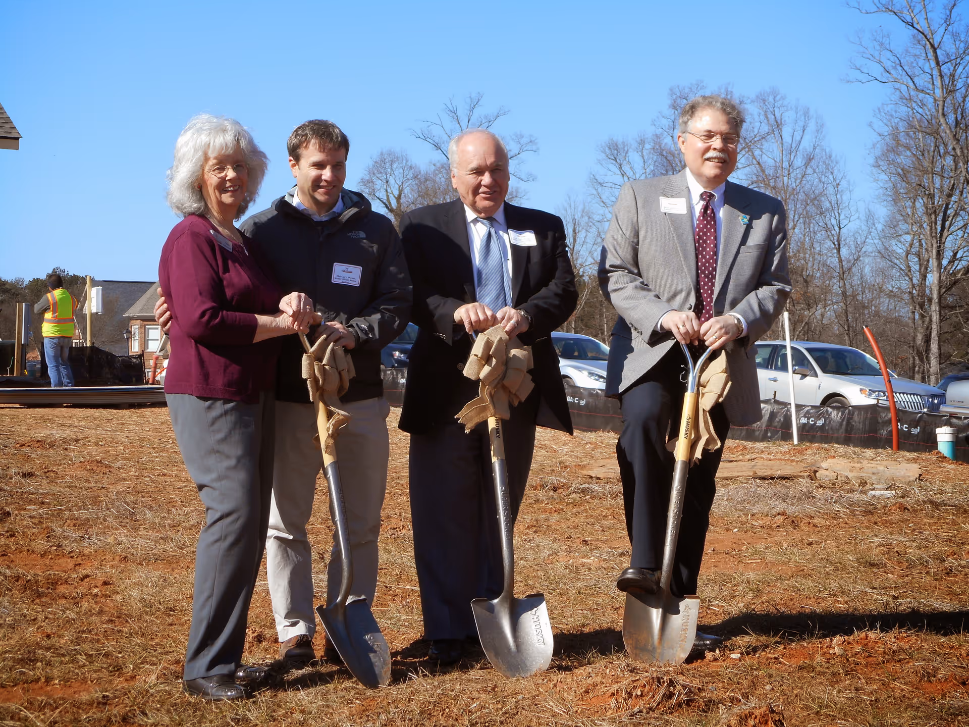 Four people standing outdoors on a dirt patch holding shovels, participating in a groundbreaking ceremony. They are dressed in business and casual attire, with cars and trees in the background under a clear blue sky.