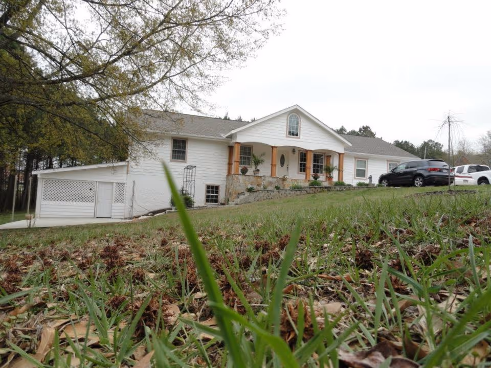 A white single-story house with a front porch supported by wooden columns, surrounded by a grassy lawn with trees in the background. Two cars are parked on the driveway to the right of the house.