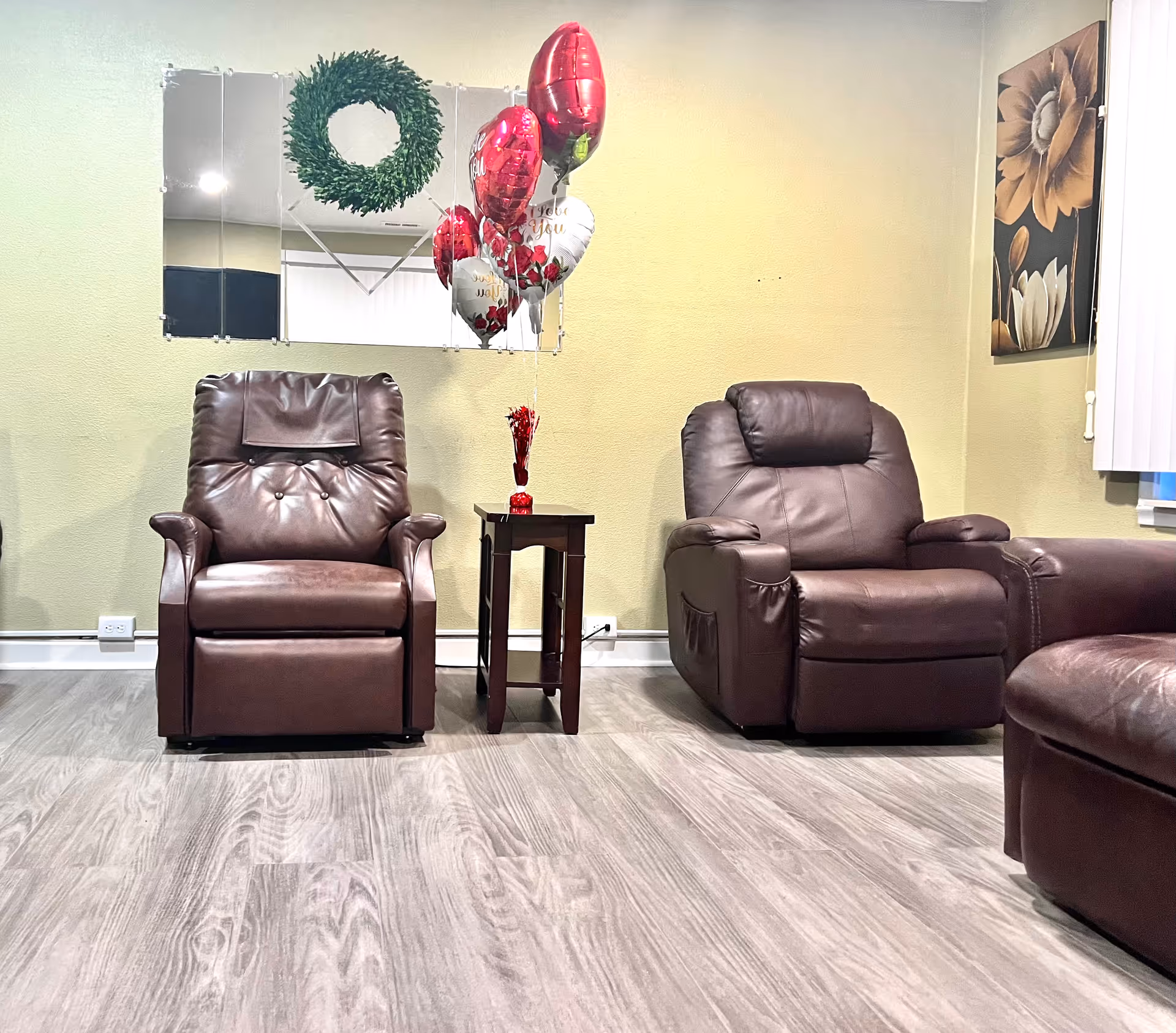 A cozy living room area with two brown leather recliners separated by a small wooden side table. Above the table, there are red and white heart-shaped balloons and a green wreath reflected in a wall mirror. The room has light-colored walls, a floral painting on the right wall, and light wood flooring.