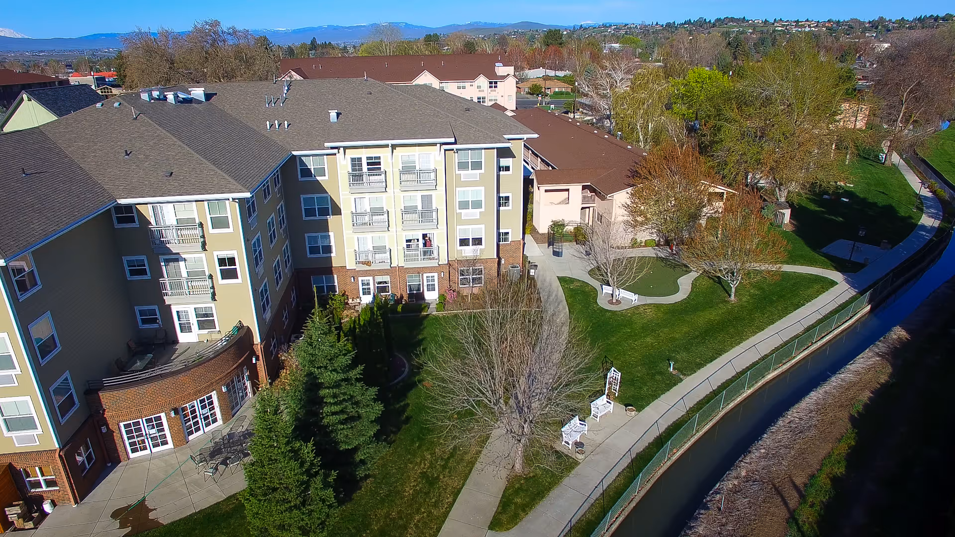 Aerial view of The Terraces at Summitview senior living facility showing a multi-story building with beige and brick exterior, surrounded by green lawns, trees, walking paths, benches, and a small water canal running alongside the property.