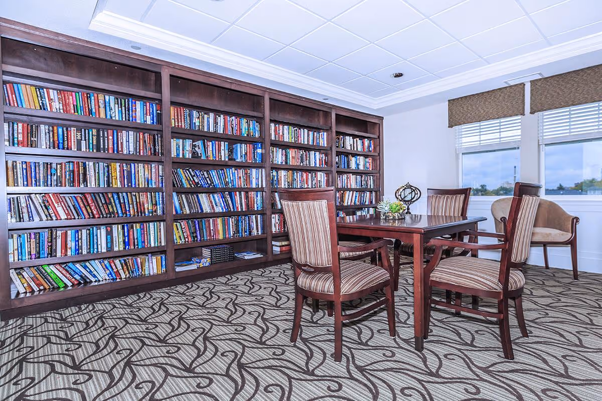 A bright room with a large wooden bookshelf filled with books along one wall. In front of the bookshelf is a wooden table with four striped upholstered chairs. There are two windows with blinds and valances letting in natural light, and a patterned carpet covers the floor.