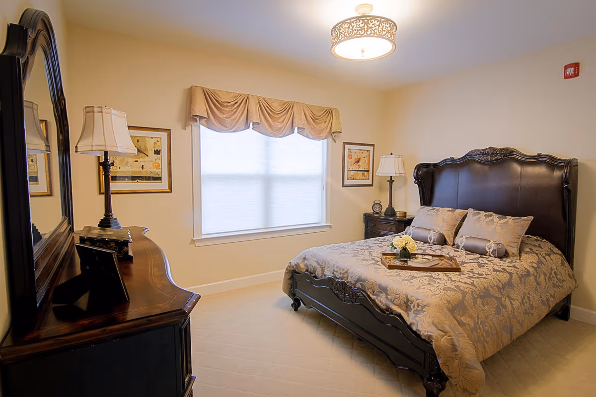 A well-lit bedroom with a large dark wooden bed featuring an ornate headboard, covered with a patterned beige and gold bedspread and matching pillows. There is a wooden dresser with a large mirror on the left side, a table lamp, and framed artwork on the walls. A window with a beige valance allows natural light into the room.