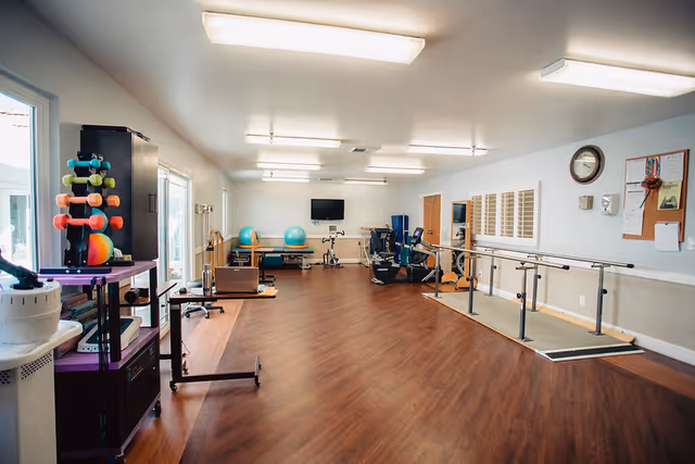 A spacious rehabilitation or physical therapy room with wooden flooring, parallel bars for walking exercises, exercise balls, stationary exercise equipment, and colorful dumbbells on a rack. The room is well-lit with ceiling lights and has large windows on one side.