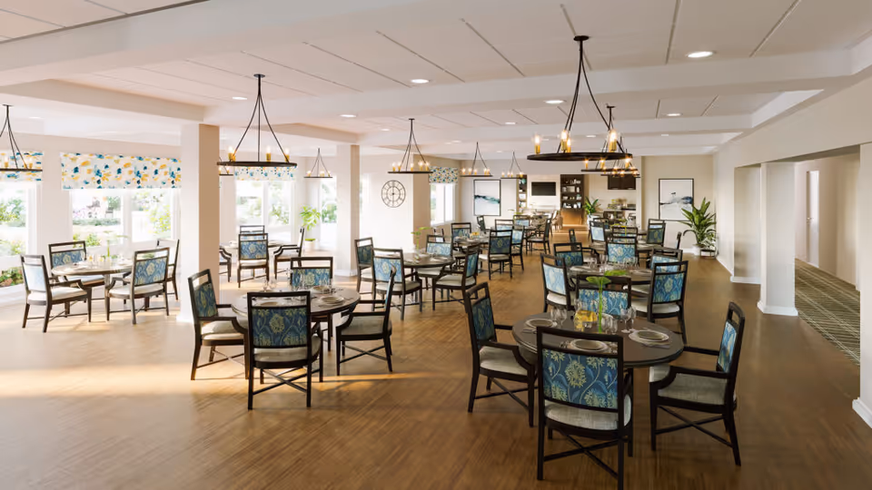 A spacious and well-lit dining room with multiple round tables and chairs arranged neatly. Each table is set with plates, glasses, and silverware. The room features large windows with floral valances allowing natural light to fill the space. Modern chandeliers hang from the ceiling, and there are decorative plants and artwork on the walls.