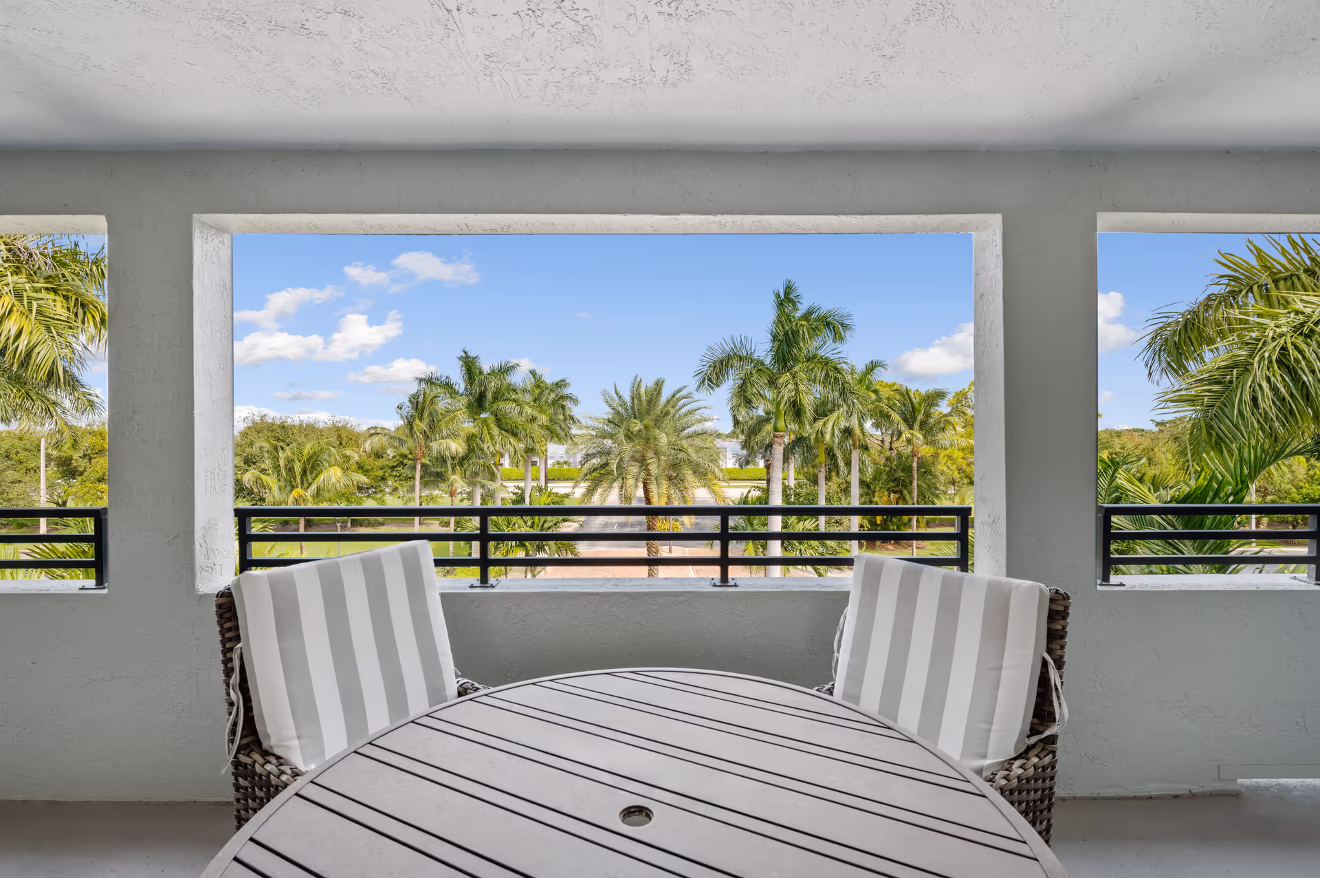 View from a covered balcony with a round table and two striped cushioned chairs, overlooking a landscape of palm trees under a blue sky with some clouds.