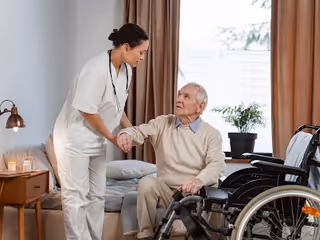 A female caregiver in white scrubs is assisting an elderly man seated in a wheelchair inside a room with a bed, a nightstand with a lamp and candles, and a potted plant near the window with brown curtains.