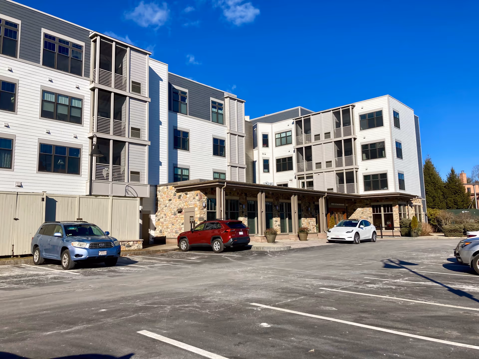 Front exterior of a multi-story senior living building with a covered entrance and several cars parked in the lot under a clear blue sky.