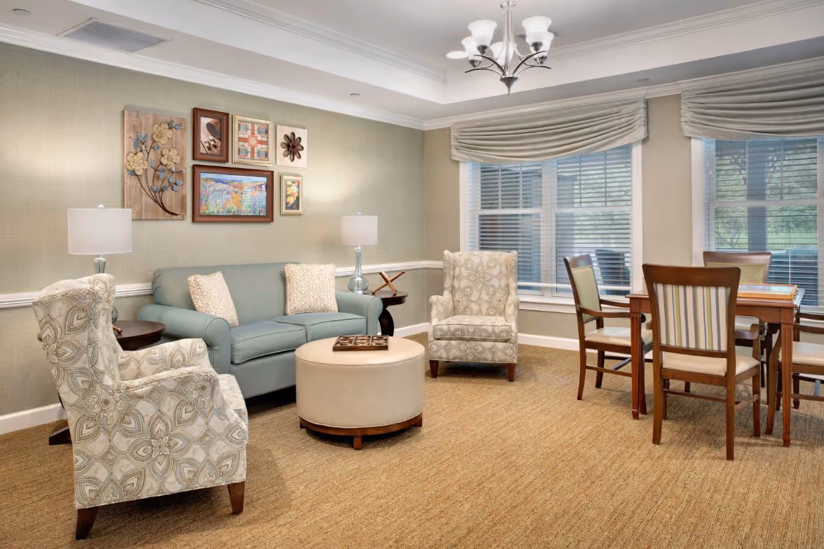 A cozy living room area with a light blue sofa, two patterned armchairs, a round beige ottoman, and a wooden table with four chairs near large windows with blinds and drapes. The walls are decorated with framed artwork and two table lamps provide soft lighting.