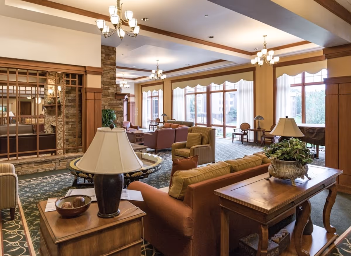 A spacious and well-lit living room area in a senior living facility with large windows allowing natural light to fill the room. The room features multiple seating arrangements including sofas and armchairs, wooden tables with lamps and decorative plants, carpeted floors with a patterned design, and chandeliers hanging from the ceiling. There is a stone fireplace visible through a wooden lattice partition on the left side.