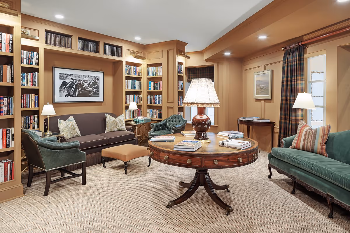 A cozy and elegant living room with built-in bookshelves filled with books, a dark brown sofa with patterned pillows, two green leather armchairs, a round wooden table with a lamp and several books on it, beige carpet, and warm beige walls with framed artwork and plaid curtains.