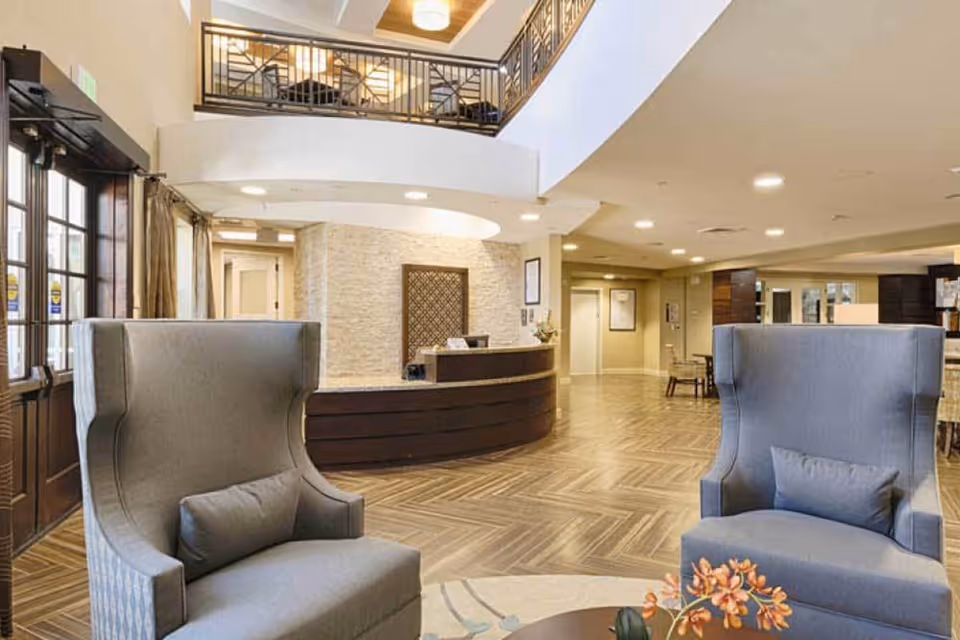 Spacious senior living facility lobby with two gray high-back armchairs and a round table with an orange flower arrangement in the foreground. The reception desk is curved and made of dark wood with a light stone wall behind it. The area features wood-patterned flooring, warm lighting, and a second-floor balcony with metal railings.